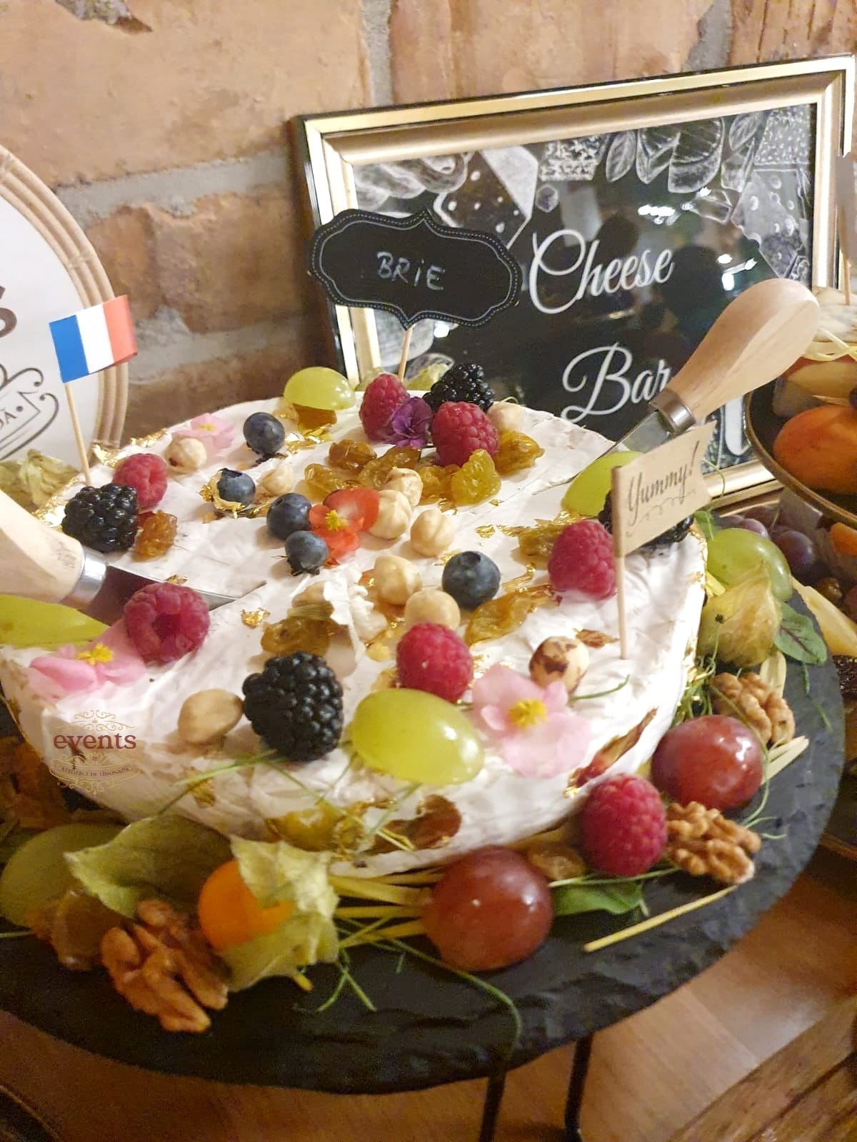 Brânză Brie rotundă decorată cu fructe de pădure, struguri, stafide, alune și flori comestibile, prezentată la Cheese Bar în fața unei tablete cu eticheta “Brie”.