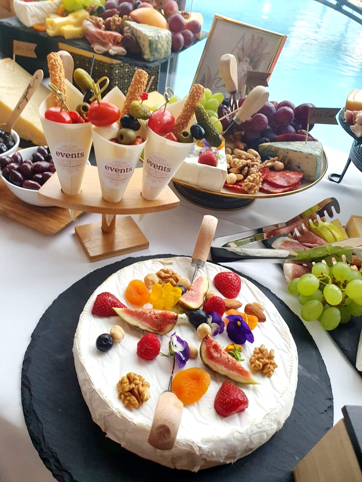 Brânză Brie decorată cu fructe, nuci și flori comestibile, alături de conuri cu grisine, măsline și roșii cherry, parte dintr-un Cheese Bar servit la un eveniment lângă piscină.