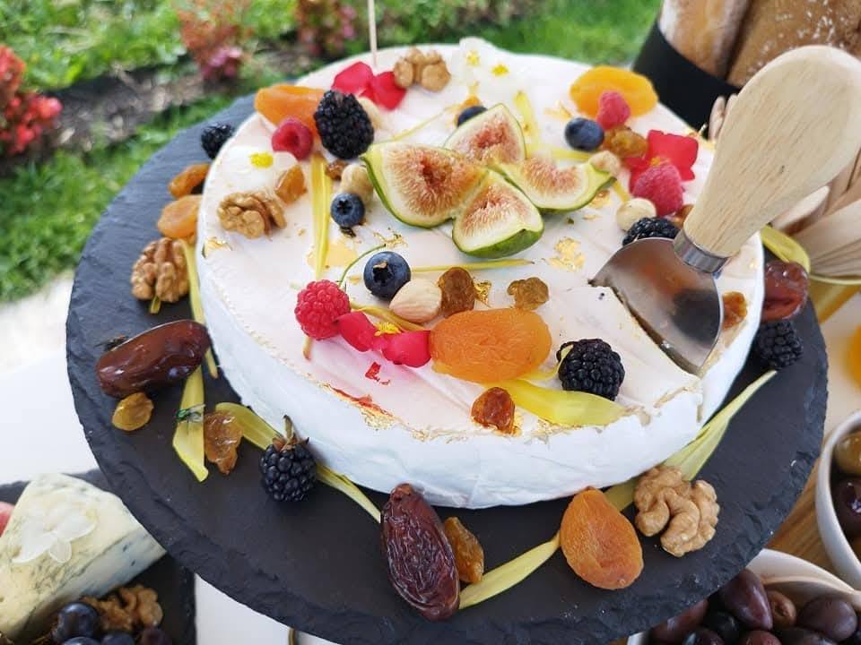 Brânză Brie decorată cu smochine, zmeură, afine, curmale, caise uscate, nuci și foiță de aur, parte dintr-un Cheese Bar servit la exterior.