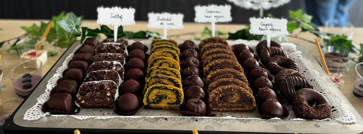 Platou cu prăjituri vegane fără zahăr, gluten, lactate sau ouă – salam de biscuiți, rulade raw, trufe și gogoși vegane din ciocolată, servite pe suport elegant.