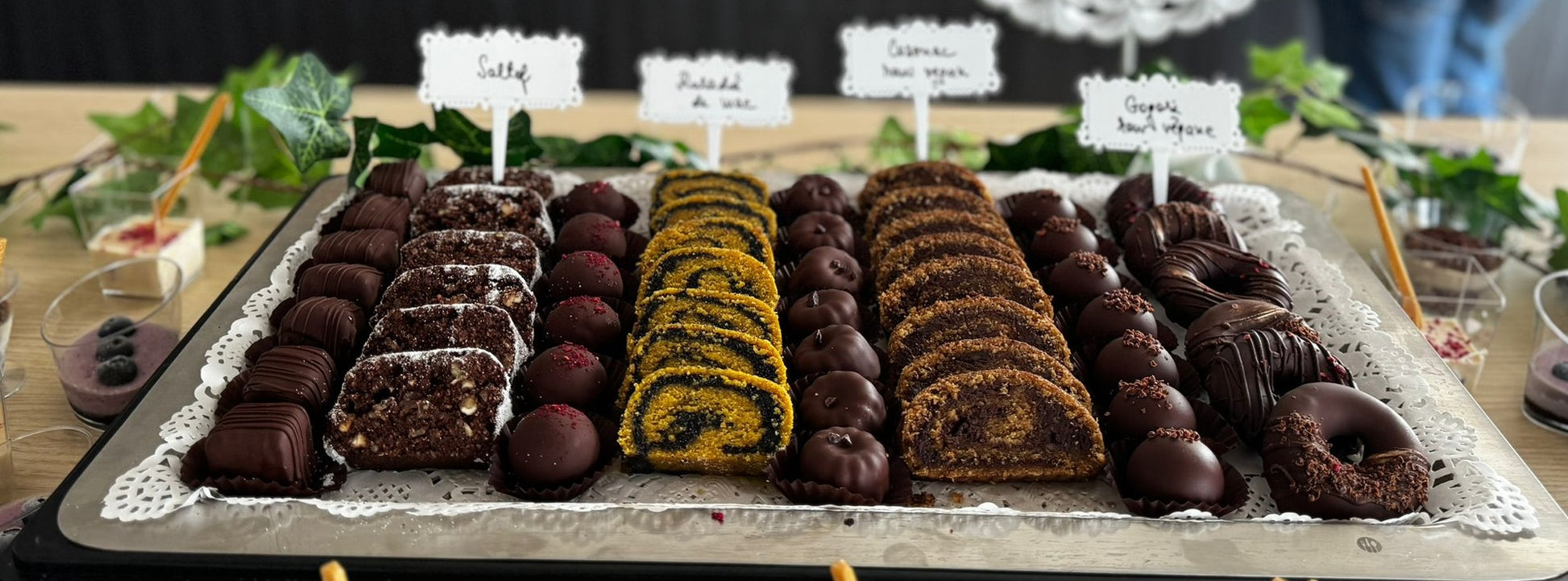 Platou cu prăjituri vegane fără zahăr, gluten, lactate sau ouă – salam de biscuiți, rulade raw, trufe și gogoși vegane din ciocolată, servite pe suport elegant.