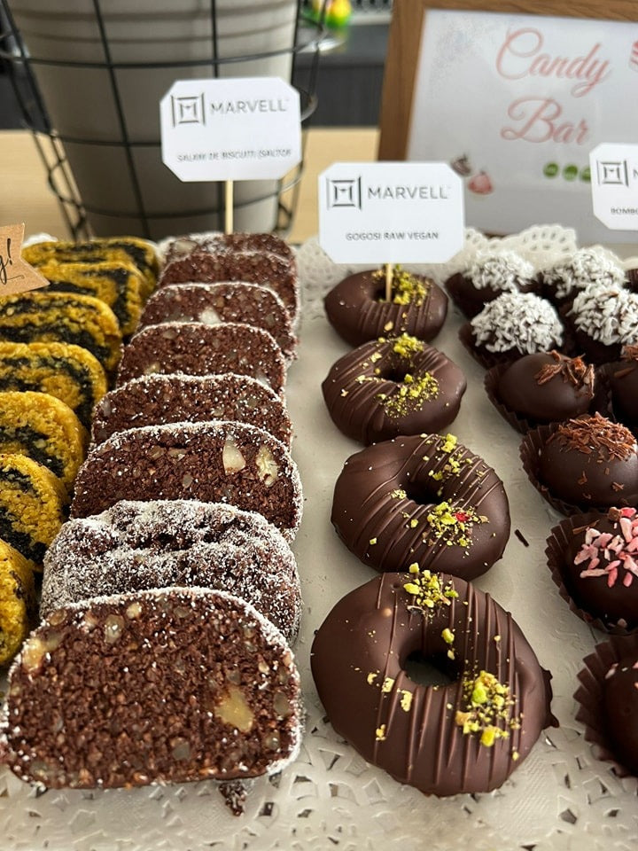 Prăjituri vegane de tip Candy Bar: salam de biscuiți raw vegan cu nuci, gogoși vegane glazurate cu ciocolată și fistic, alături de bomboane decorate cu fulgi de cocos, ideale pentru evenimente fără zahăr, gluten sau lactate.