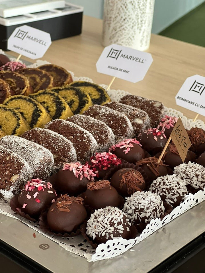 Ruladă vegană cu mac și cacao, alături de bomboane vegane decorate cu fulgi de cocos și ciocolată, fără zahăr, gluten, lactate sau ou, servite elegant pe platou.