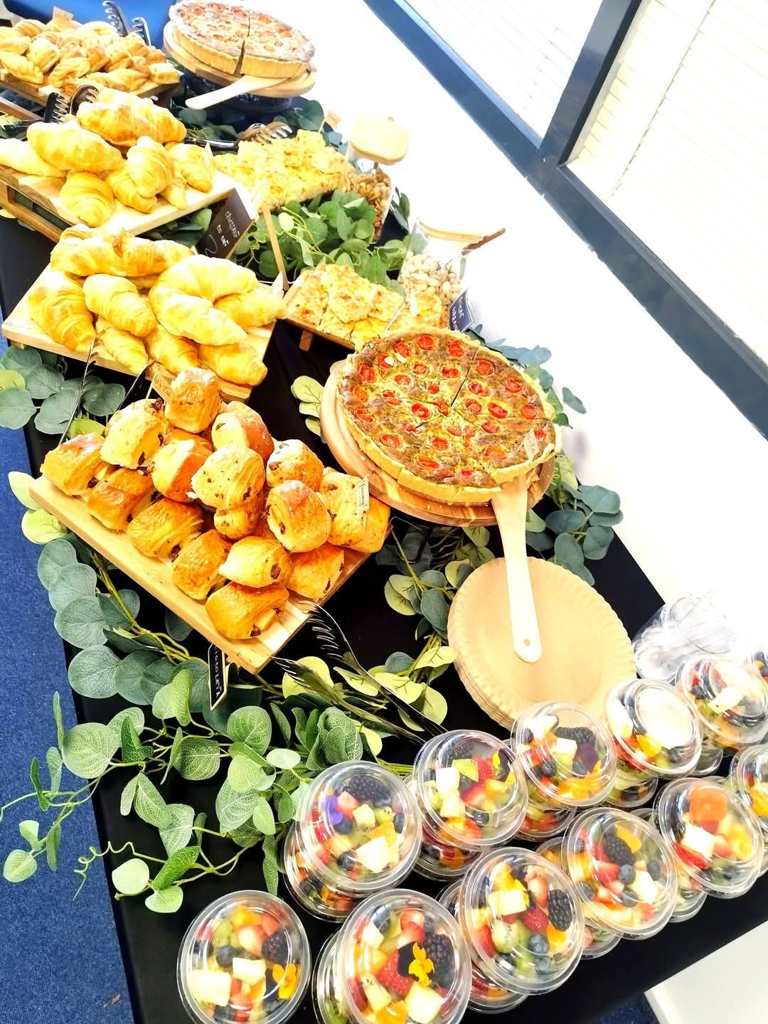Masă de catering pentru pauza de cafea la evenimente corporate, cu salate de fructe la caserolă, croissante, pateuri cu ciocolată și quiche-uri.