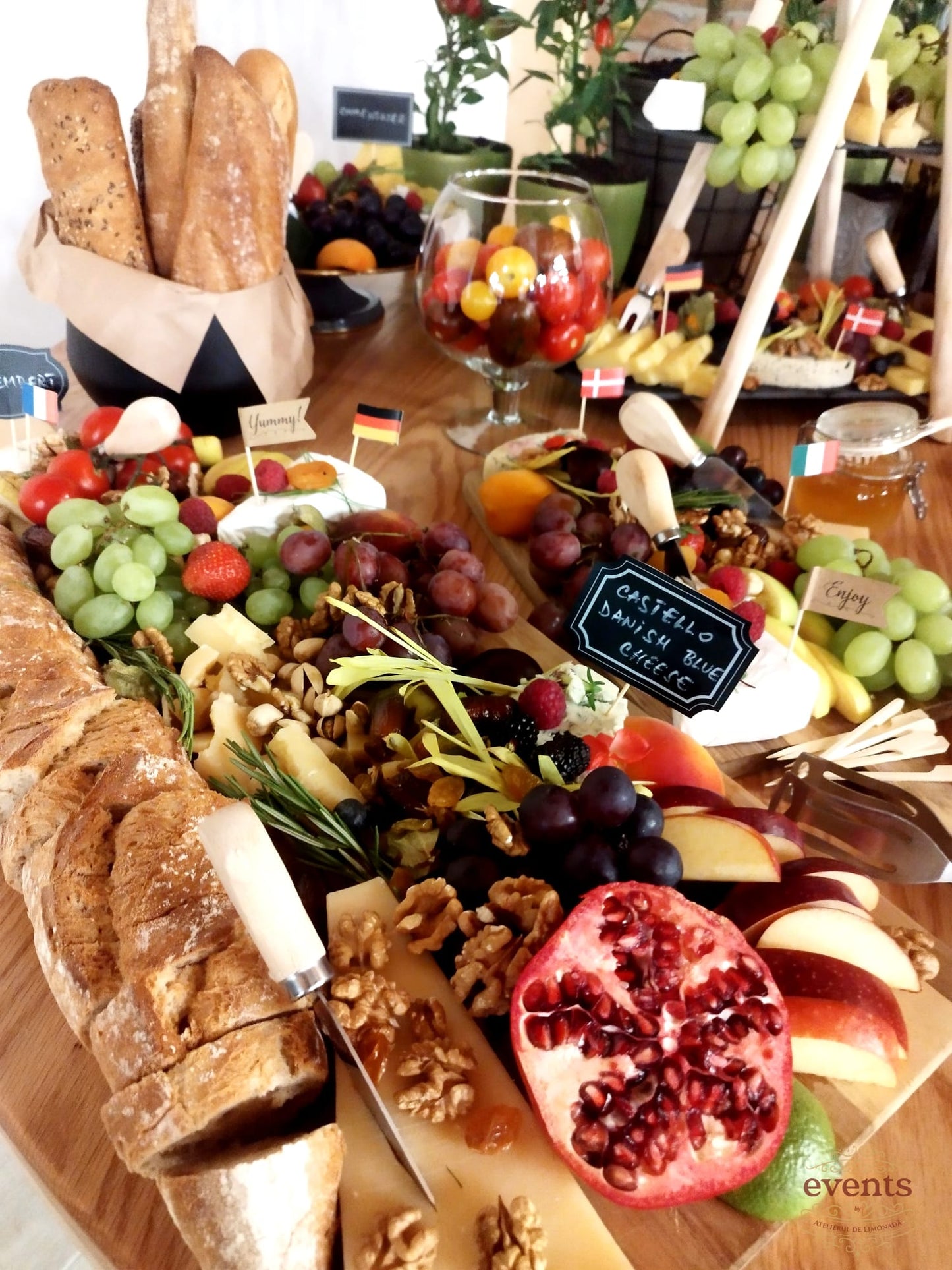 Cheese Bar bogat cu brânzeturi, struguri, nuci, rodie, pâine artizanală, steaguri tematice și borcan cu roșii cherry, pe fundal rustic cu decor vegetal.