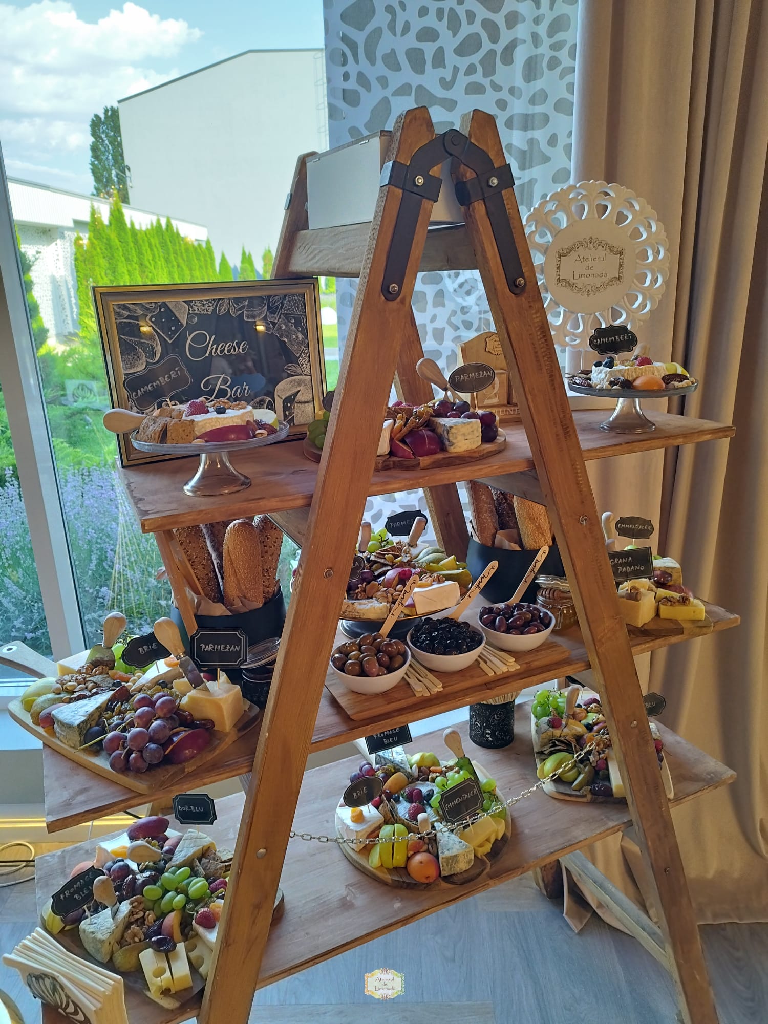 Cheese Bar creativ, aranjat pe un suport din lemn tip scară, cu platouri de brânzeturi, struguri, măsline, pâini artizanale și decor personalizat cu etichete.