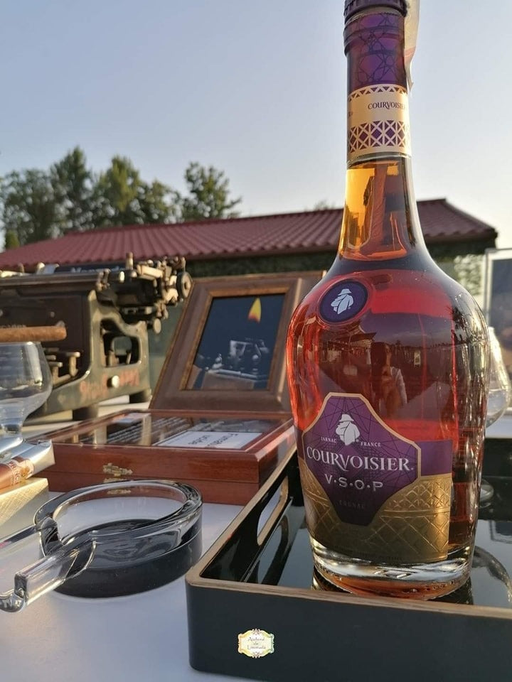 Sticlă de cognac Courvoisier alături de trabucuri, scrumieră de sticlă și elemente vintage pentru un cigar bar elegant în aer liber.