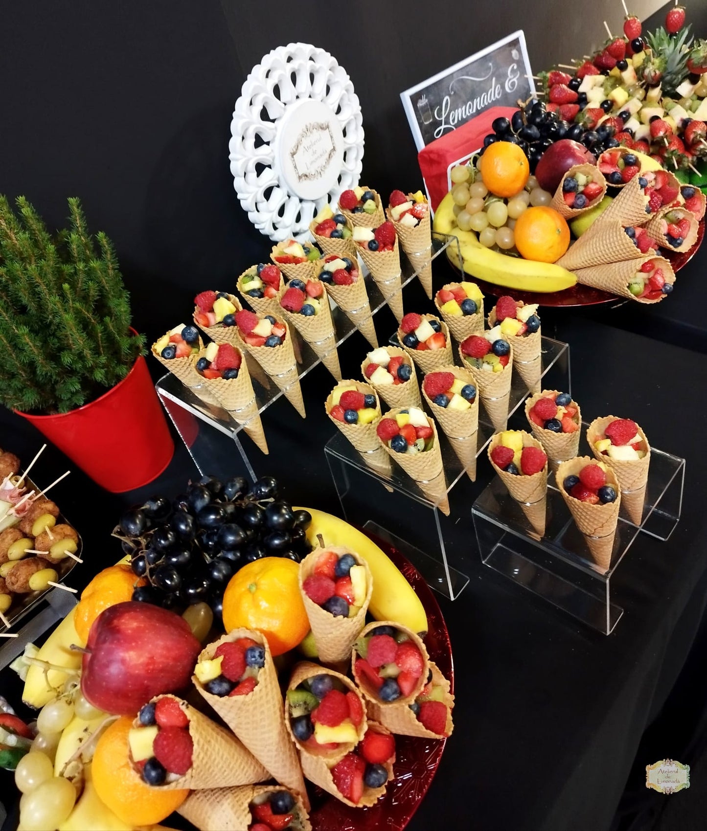 Conuri cu fructe proaspete – afine, zmeură, ananas, struguri – aranjate pe suporturi transparente într-un fruit bar elegant, alături de fructe întregi și aranjamente tematice.