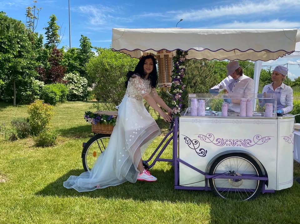 Mireasă alături de o tricicletă albă decorată, transformată în ice cream bar cu gelato artizanal pentru nunți.