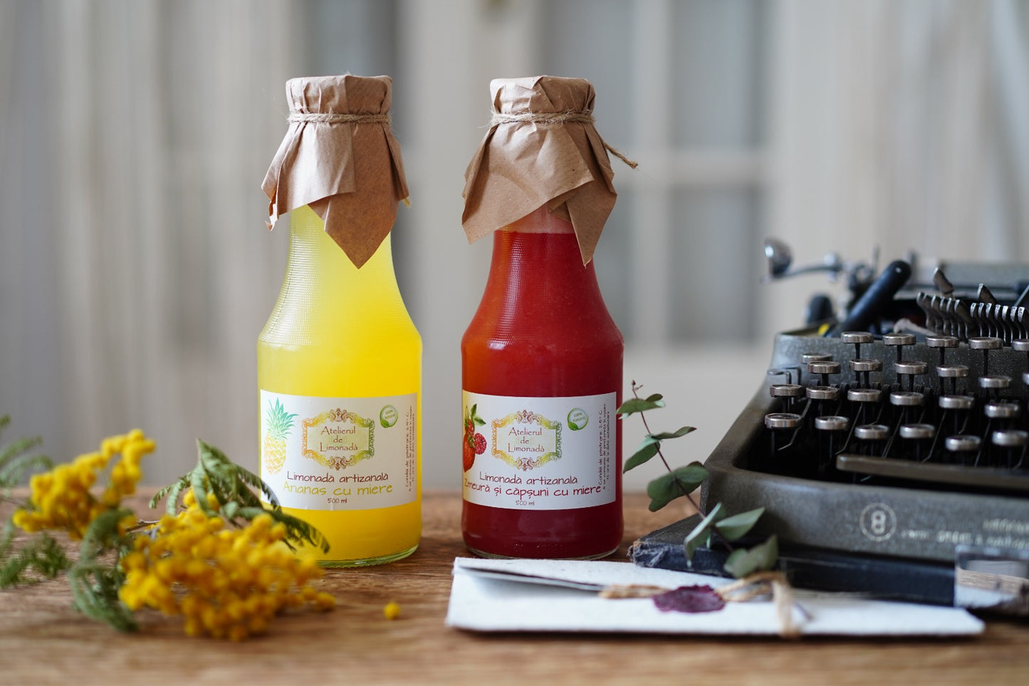 Două sticle de limonadă la sticlă, cu ananas și cu zmeură și căpșuni, așezate pe o masă de lemn lângă o mașină de scris vintage, flori galbene și o scrisoare sigilată.