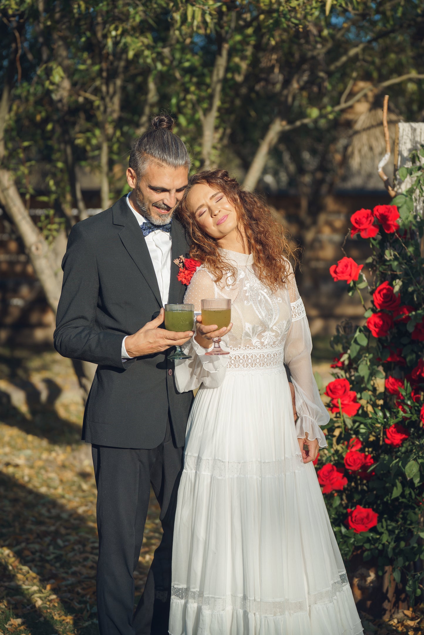Cuplu de miri savurând limonadă naturală în grădină, lângă un decor cu trandafiri roșii, într-un cadru romantic de nuntă.