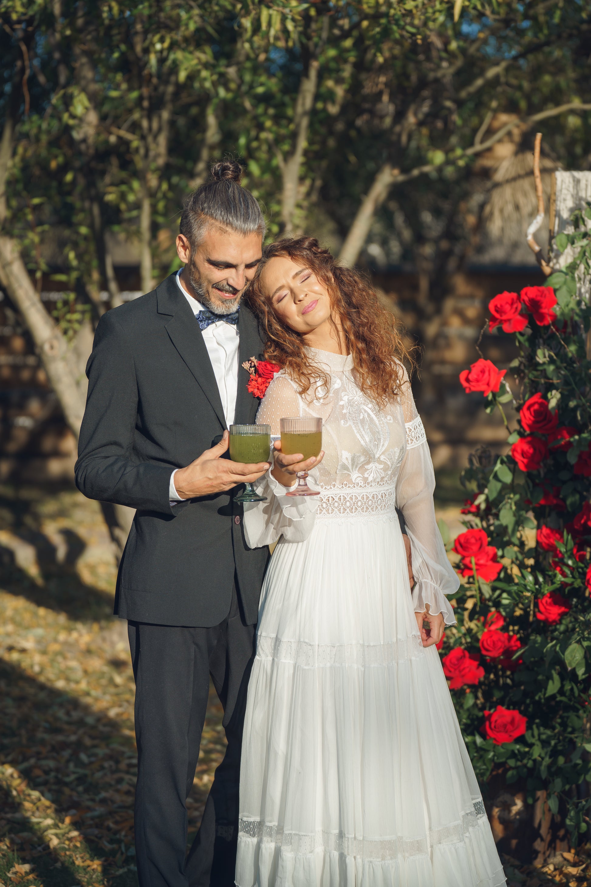 Cuplu de miri savurând limonadă naturală în grădină, lângă un decor cu trandafiri roșii, într-un cadru romantic de nuntă.
