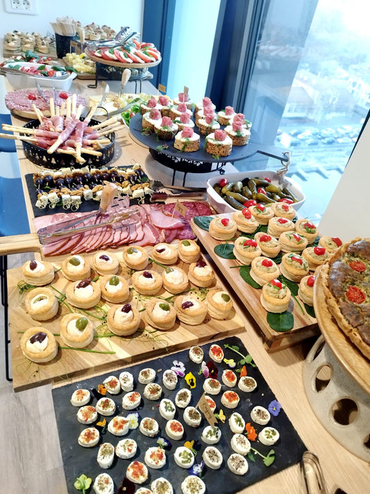 Bufet cu mix aperitive pentru eveniment, incluzând grisine cu jamon, brânzeturi, rulouri, tartine, mini-foietaje, ouă umplute, mezeluri, castraveți murați și roșii cherry.