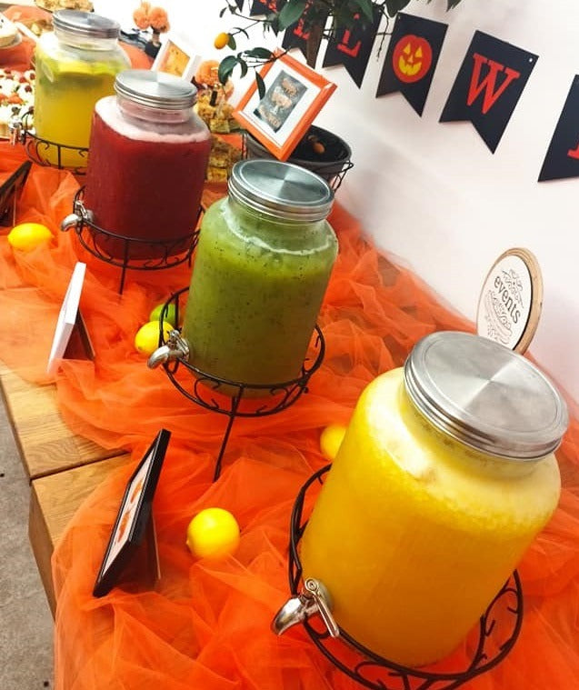 Smoothie bar cu băuturi naturale colorate servite în dispensere, decor tematic de Halloween cu fructe proaspete și elemente portocalii.
