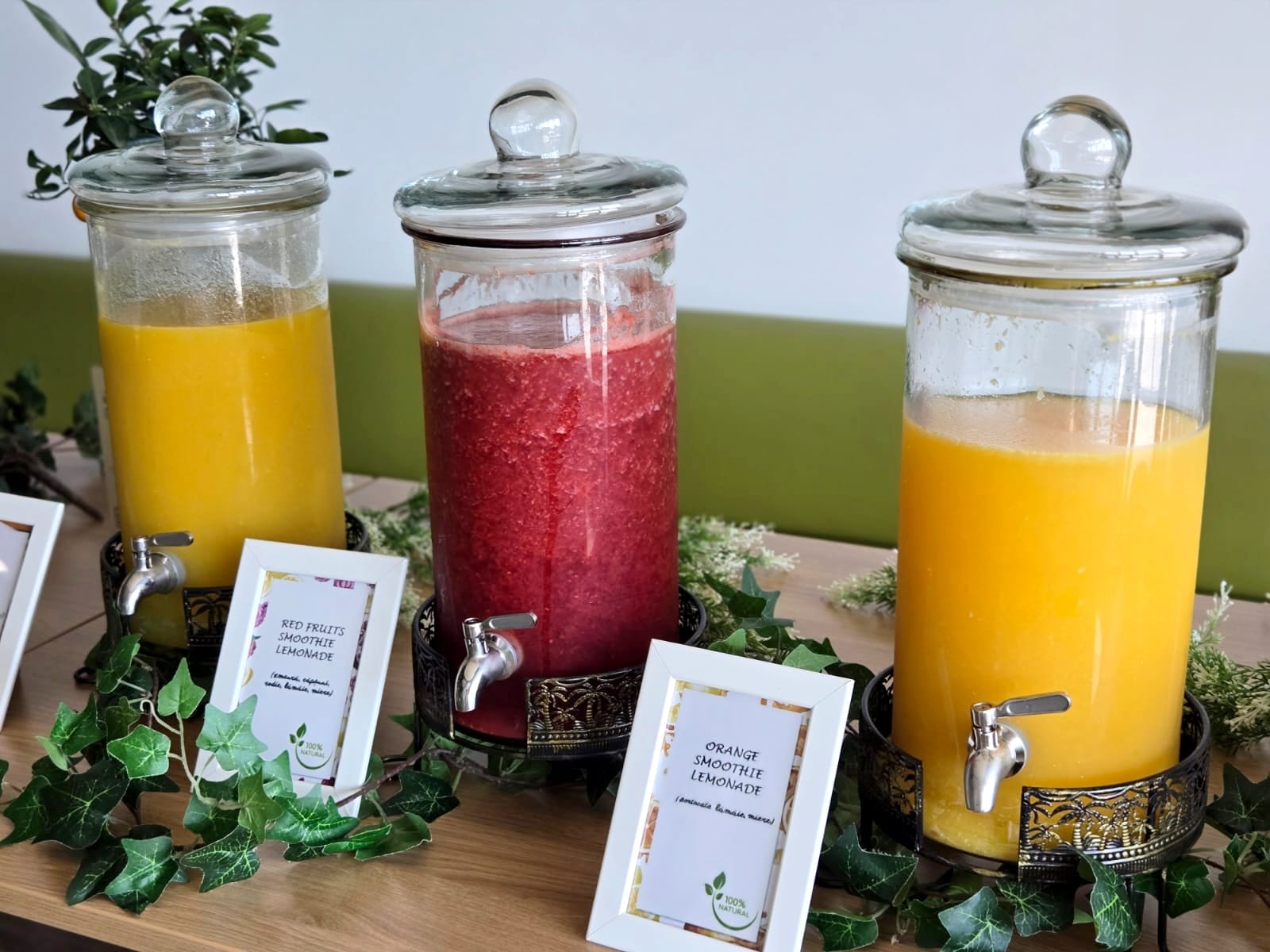 Close-up cu sortimente de smoothie: Red Fruits Smoothie Lemonade și Orange Smoothie Lemonade. Băuturi proaspete și naturale în dispensere de sticlă pe suporturi metalice. | Atelierul de Limonadă