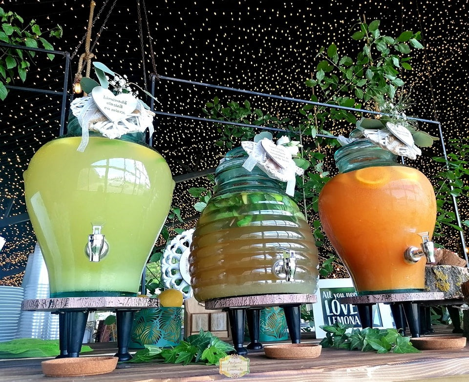 Stand de limonadă cu decor verde și lumini ambientale, conținând sortimente de limonadă clasică, cu mentă și cu portocale, servite în recipiente elegante.
