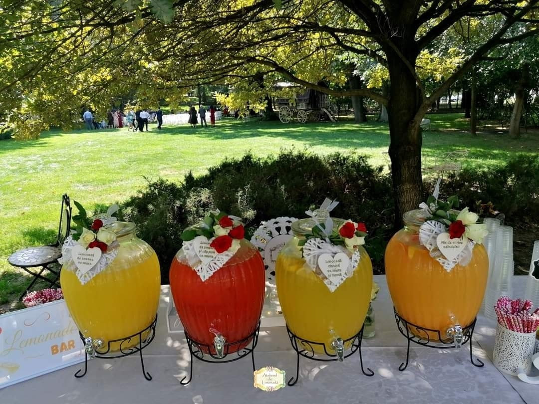 Stand de limonadă în aer liber, cu patru sortimente colorate și decor floral, la un eveniment elegant.
