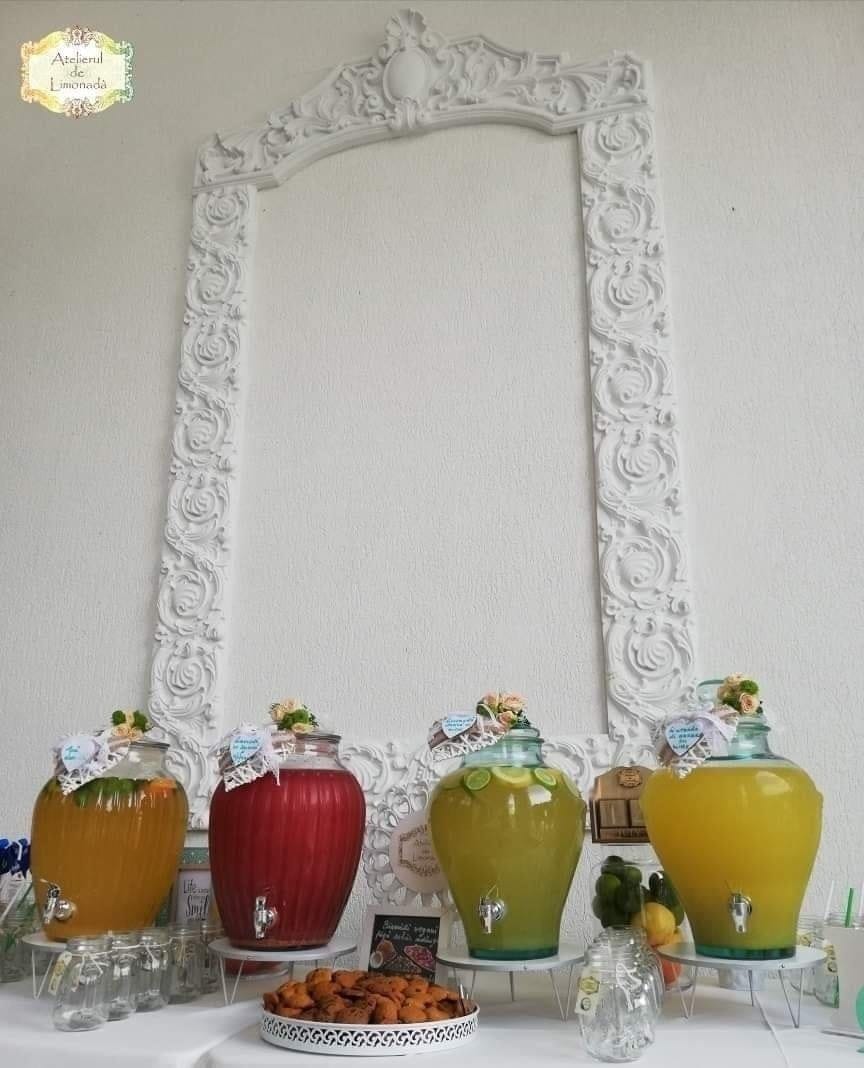 Stand de limonadă elegant cu patru sortimente colorate, decorat cu fructe și dulciuri, amplasat sub un cadru baroc alb, perfect pentru evenimente rafinate.