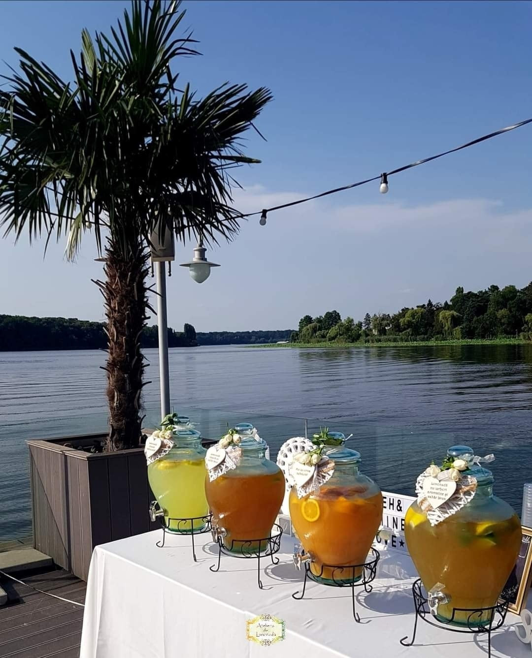 Stand de limonadă pe malul apei, cu patru sortimente colorate, decorat pentru un eveniment estival pe terasă, sub palmier.