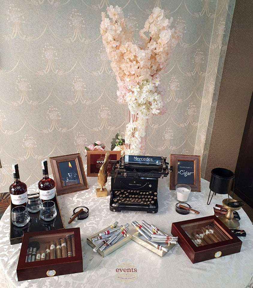 Gentleman's corner cu trabucuri premium, cognac de colecție, pahare elegante, decor floral și mașină de scris vintage pentru evenimente rafinate.