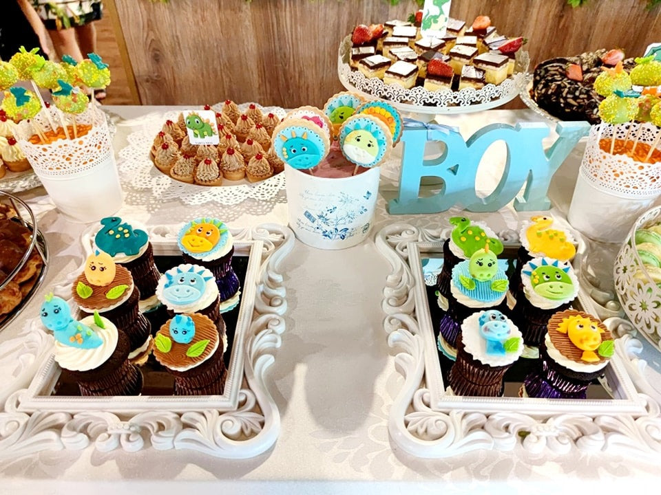 Candy bar pentru botez băiat, decorat cu cupcakes tematice dinozauri, prăjituri asortate și elemente decorative albastre, realizat de Atelierul de Limonadă.