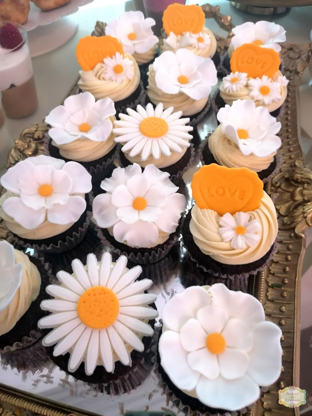 Brioșe decorate cu flori comestibile tip margarete și mesaje "love", aranjate pe un platou decorativ din candy bar-ul Atelierului de Limonadă.