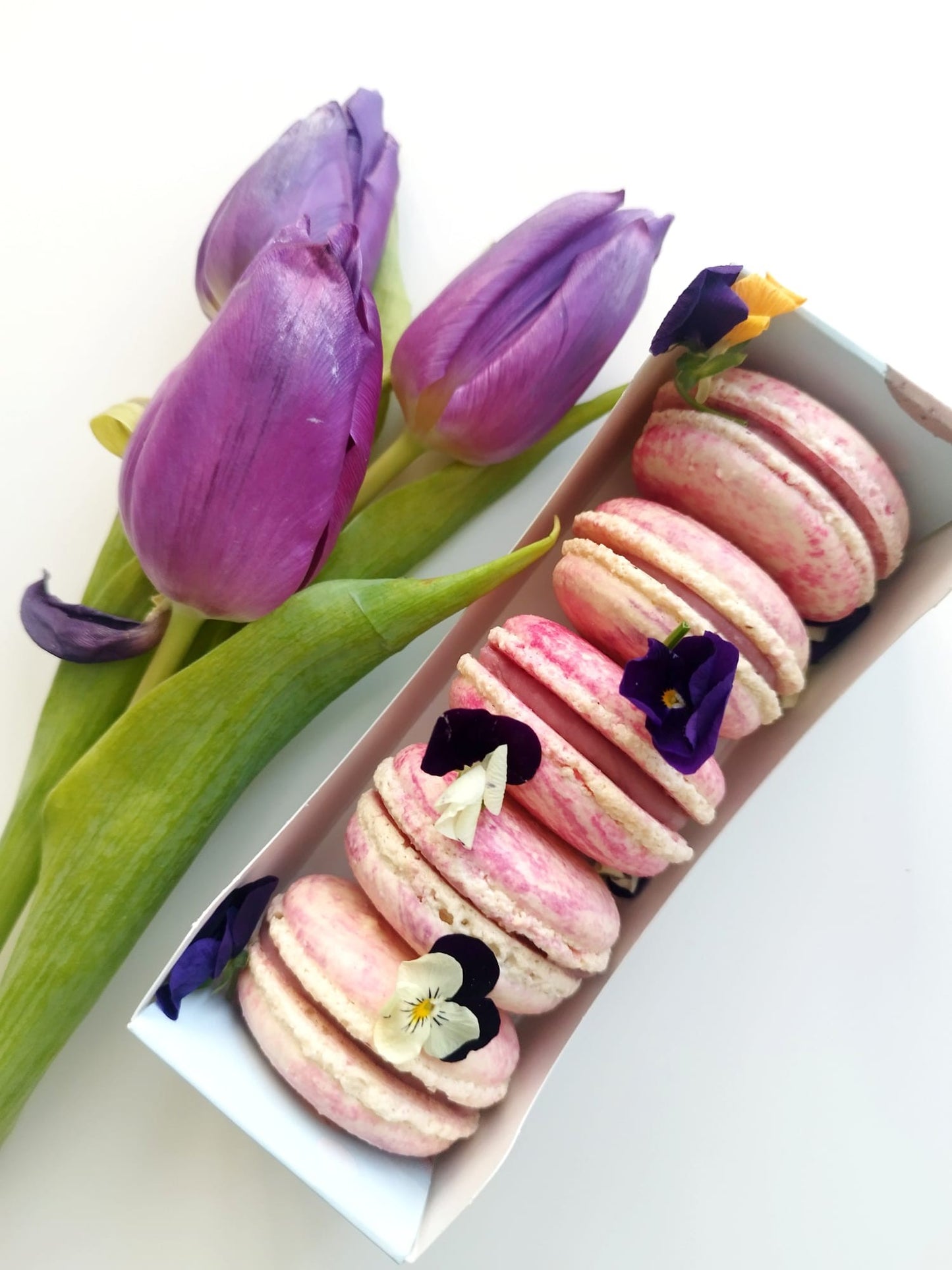 Cutie cadou cu macarons roz marmorat, decorați cu panseluțe comestibile, alături de un buchet de lalele mov.