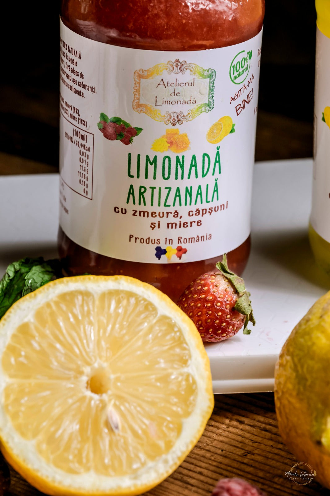 Detaliu etichetă limonadă artizanală cu zmeură, căpșuni și miere, lângă o lămâie proaspătă.