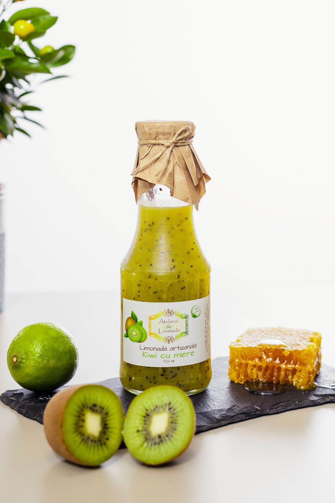 Sticlă de limonadă la sticlă cu kiwi și miere, însoțită de felii de kiwi, lime și fagure natural, pe fundal alb.