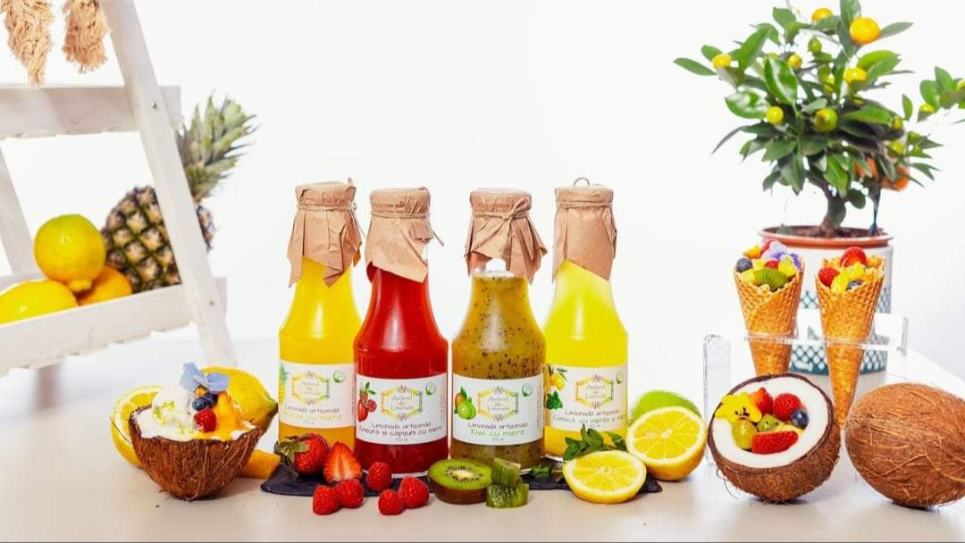 Limonadă la sticlă în patru sortimente: mango, căpșuni și zmeură, kiwi, și mentă cu lime, prezentate alături de fructe proaspete și conuri cu fructe.