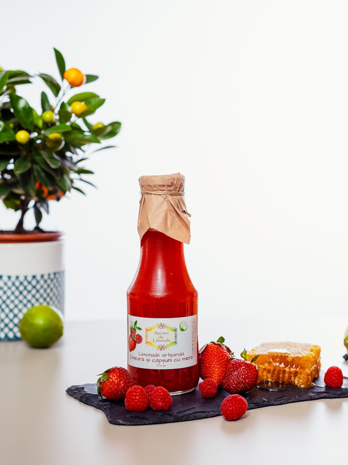 Sticlă de limonadă cu zmeură, căpșuni și miere, alături de fructe proaspete și fagure natural, pe fundal alb decorat minimalist.