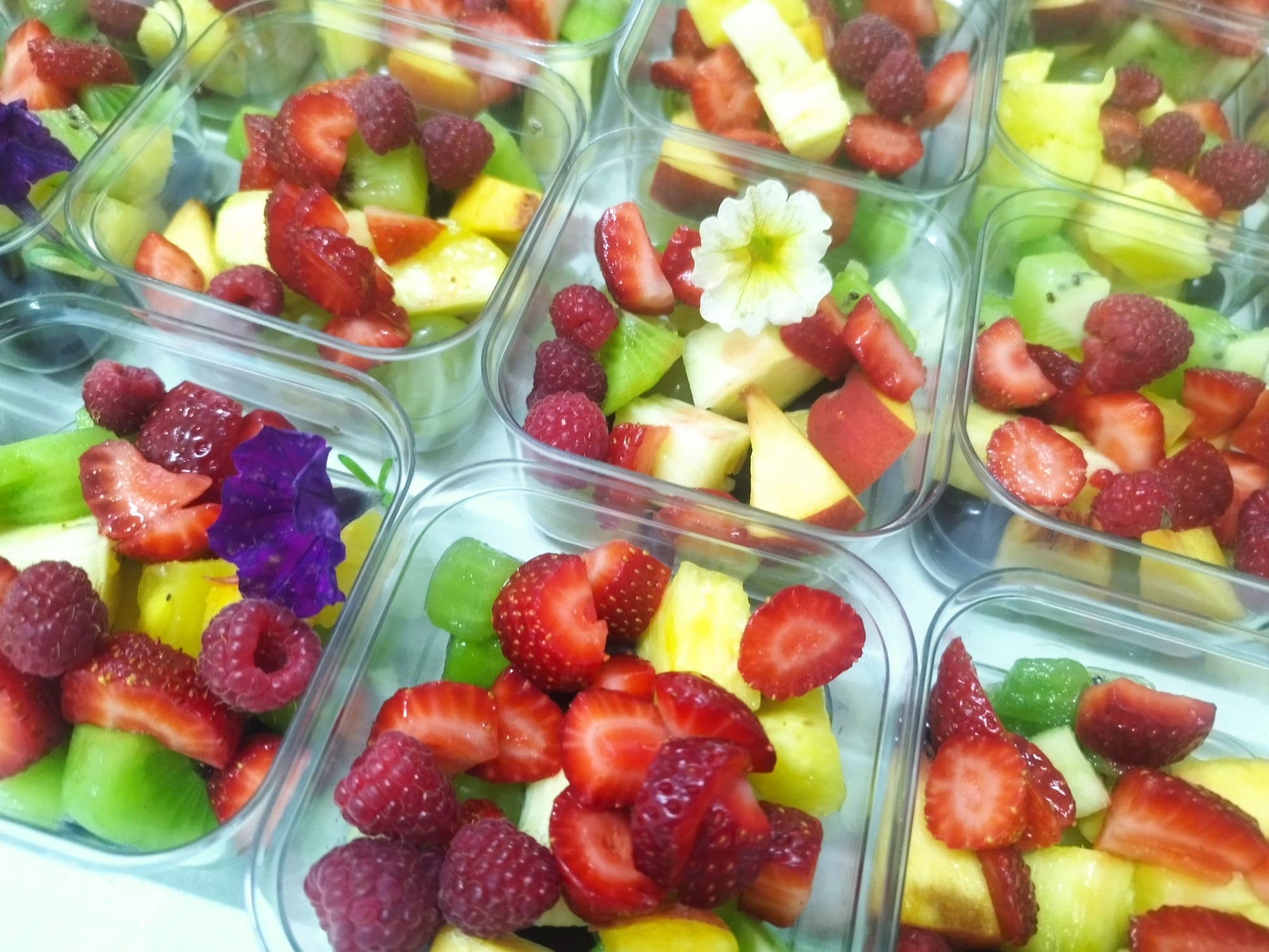 Detalii meniu catering corporate: caserole individuale cu mix de fructe proaspete, incluzând căpșuni, zmeură, kiwi și ananas, decorate cu flori comestibile.