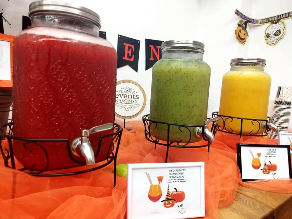 Smoothie bar decorat pentru Halloween, cu băuturi naturale în dispensere – smoothie de fructe roșii, kiwi și portocale
