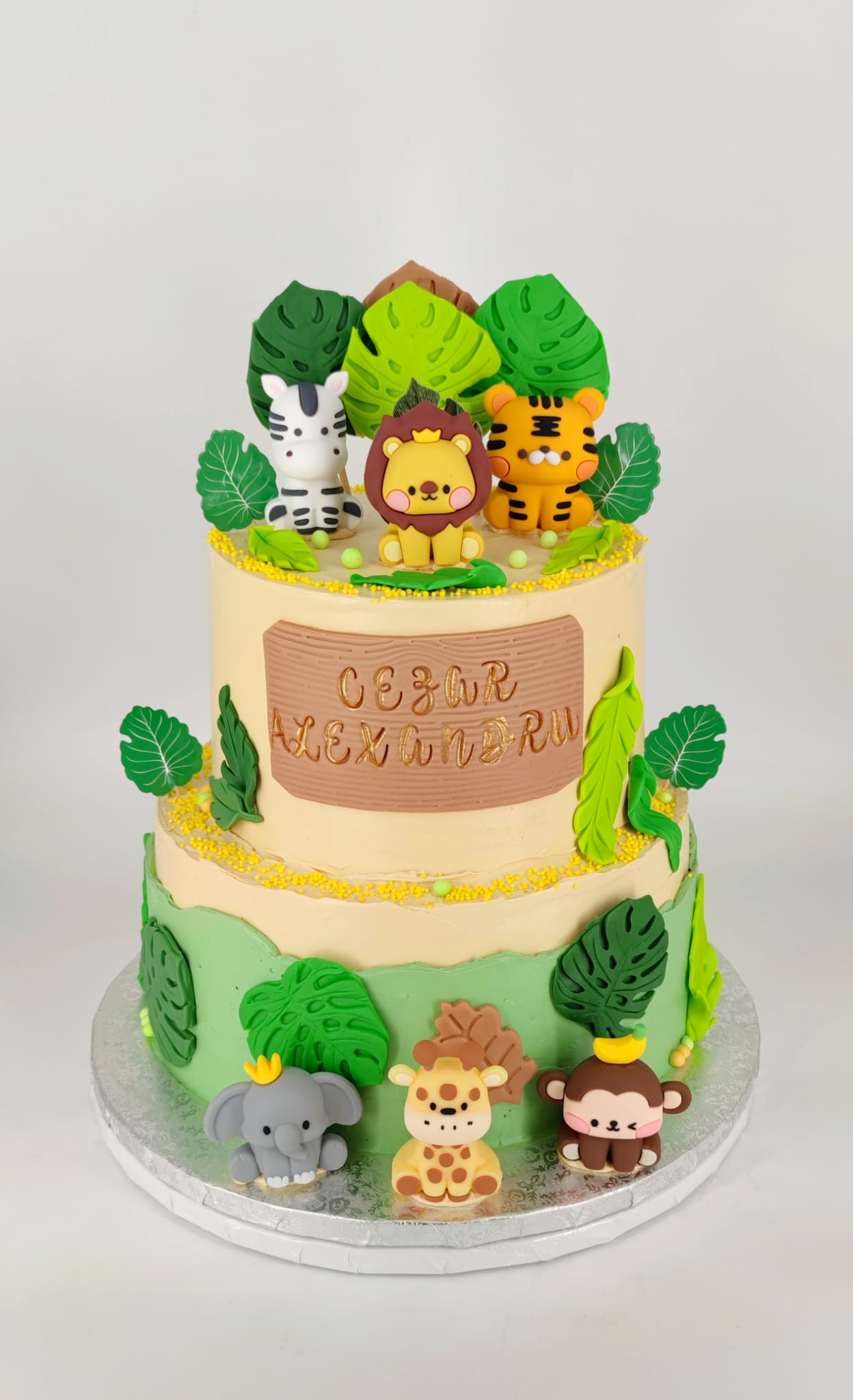 Tort de aniversare etajat, cu tematică safari, personalizat cu numele Cezar Alexandru și decorat cu figurine drăgălașe: leu, tigru, zebră, elefant și maimuță.