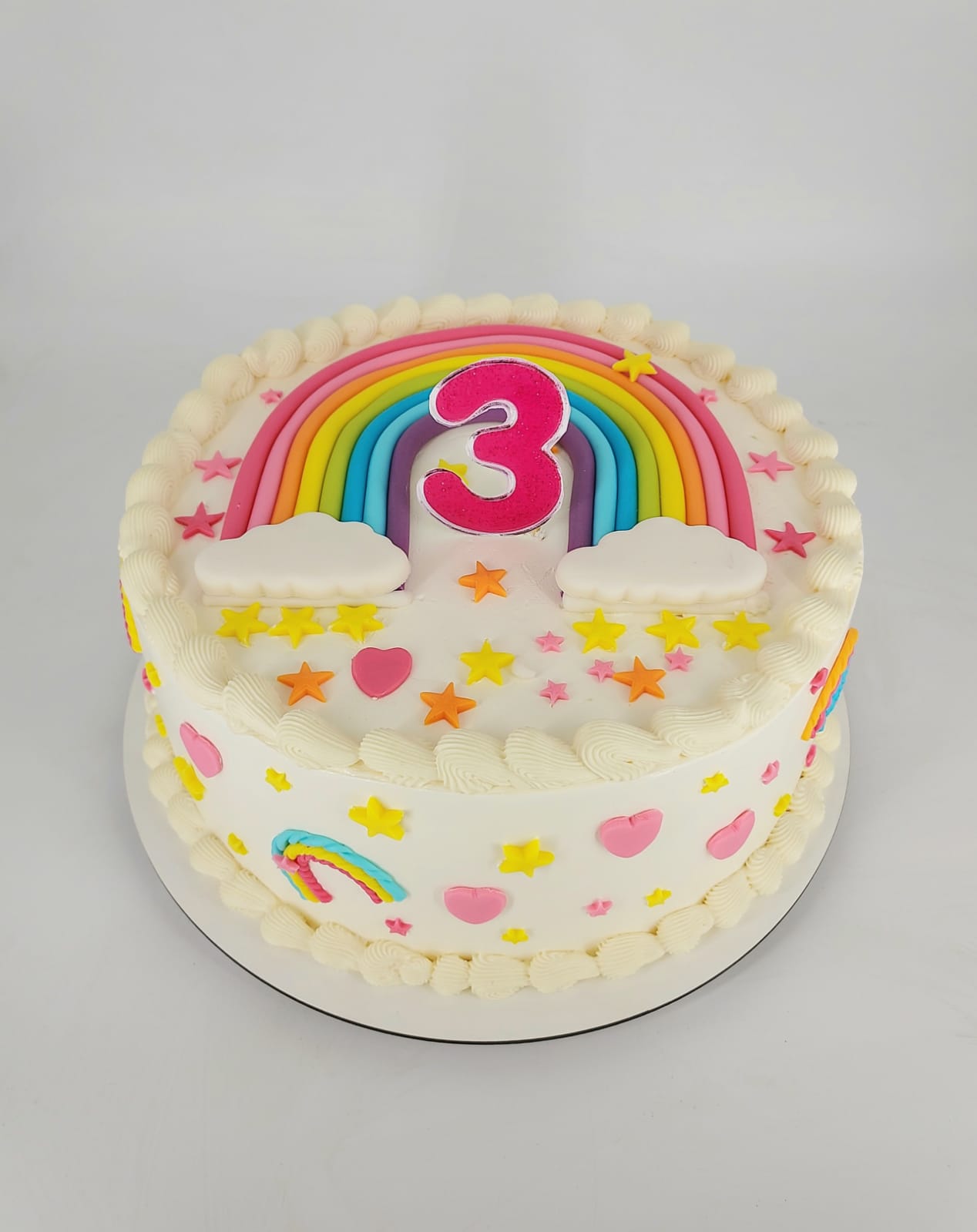 Tort rotund pentru copii, cu glazură albă, decorat cu un curcubeu mare, cifra 3 roz sclipitoare și norișori din zahăr.