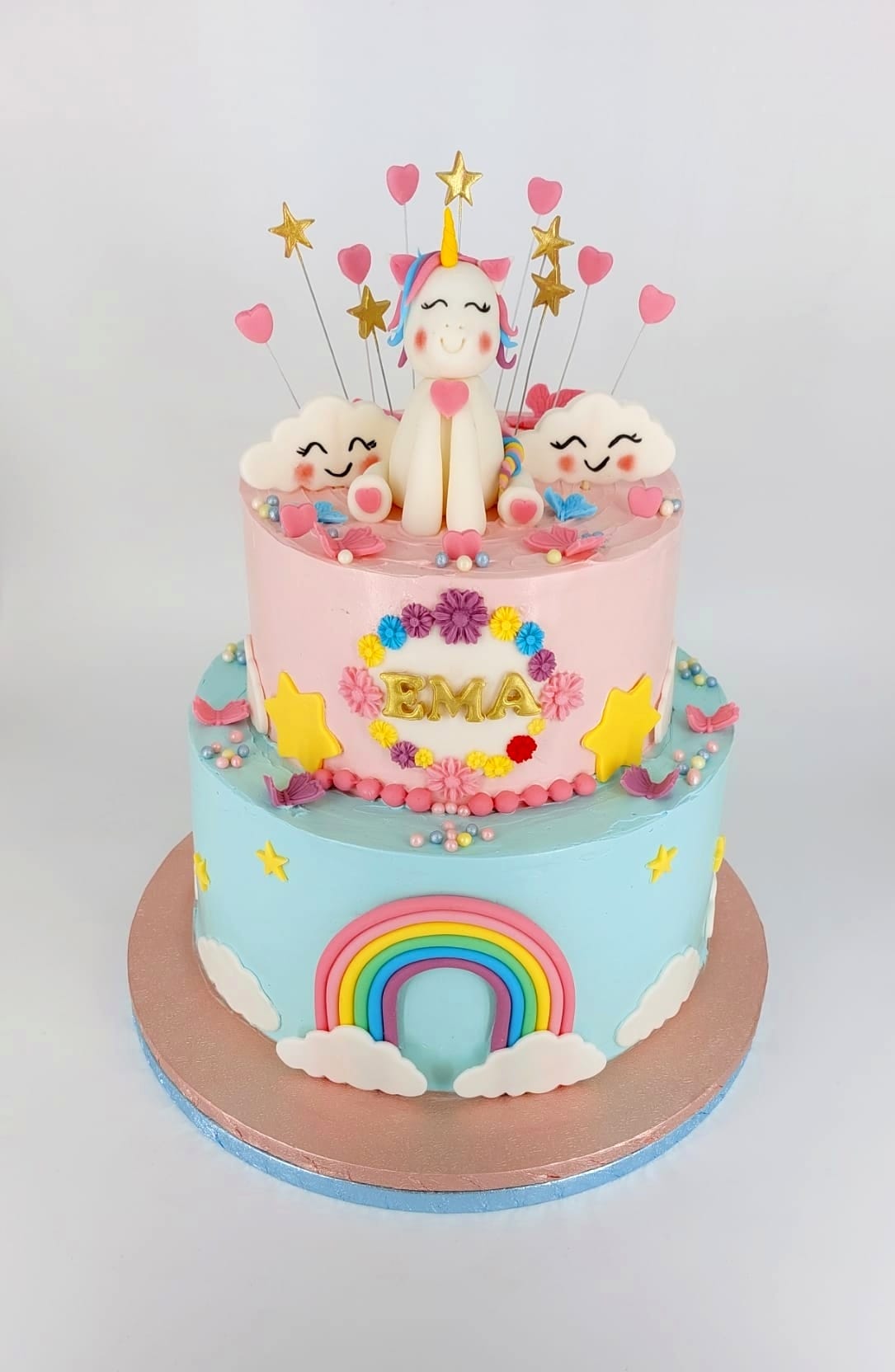 Tort etajat pentru fetițe, roz și bleu, cu un unicorn simpatic, norișori zâmbitori și un curcubeu, personalizat cu numele Ema.
