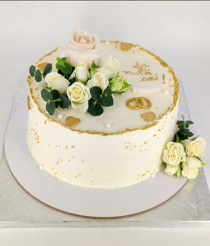 Tort alb elegant pentru aniversarea căsătoriei, decorat cu trandafiri naturali, verighete aurii și stropi de aur.