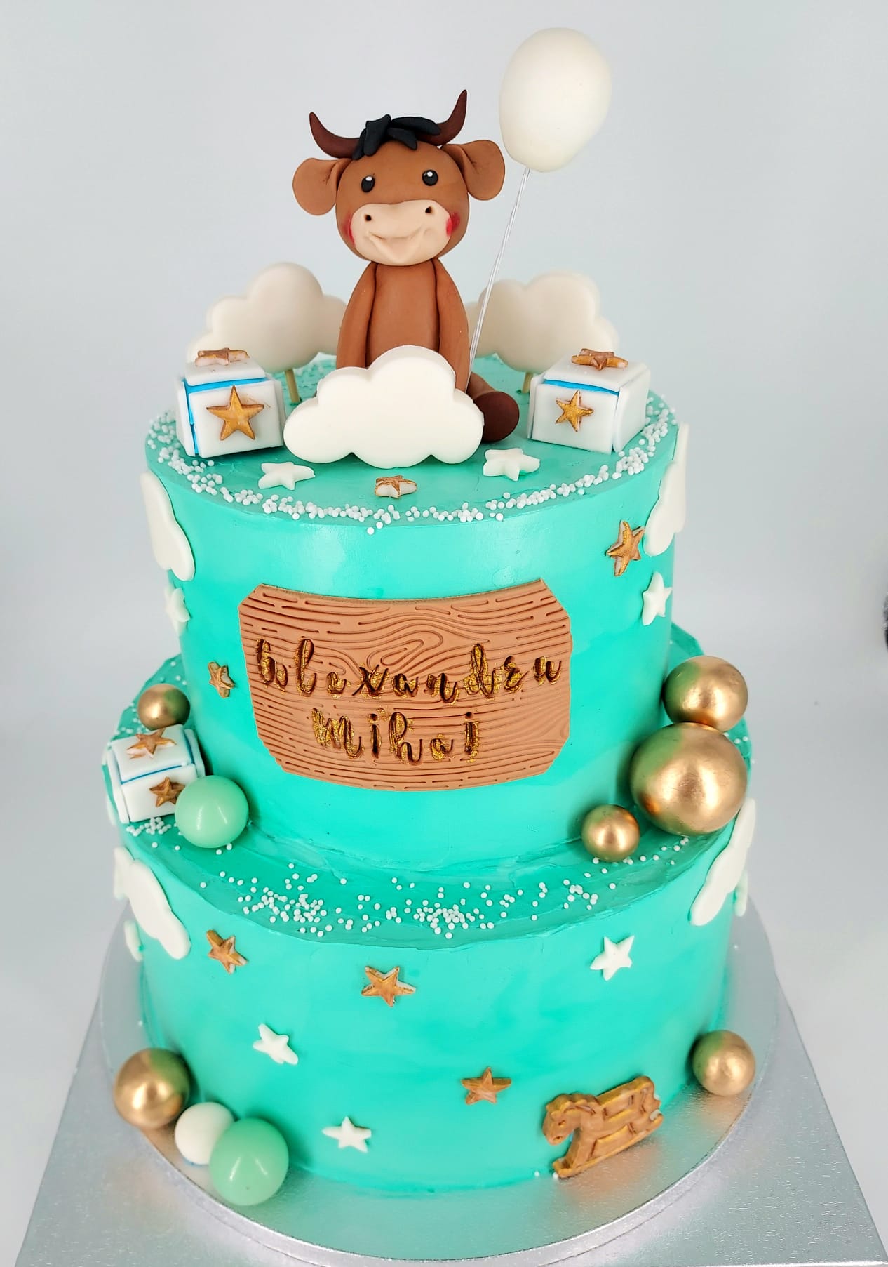 Tort de botez etajat, de culoare turcoaz, pentru băieței, personalizat cu numele Alexandru Mihai și decorat cu o figurină văcuță, norișori și steluțe aurii.