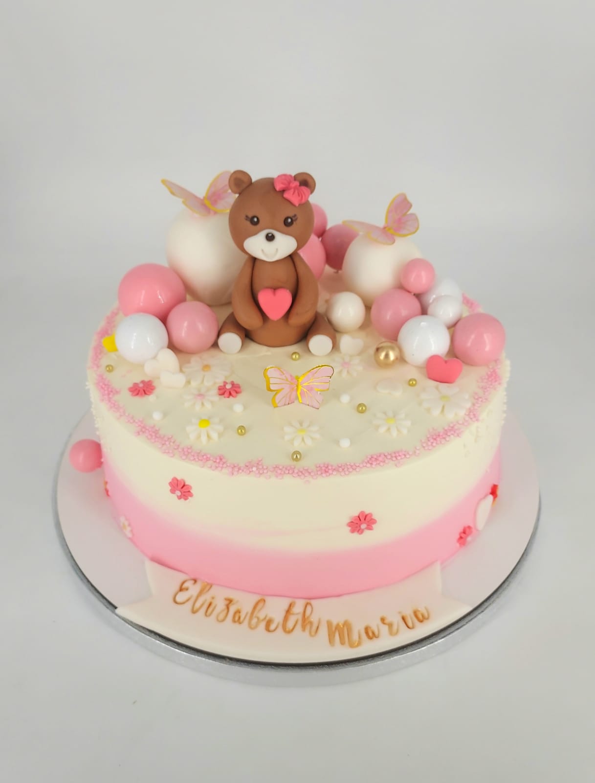 Tort personalizat de botez pentru fetițe, decorat cu un ursuleț din zahăr, buline comestibile roz și albe, și flori delicate.