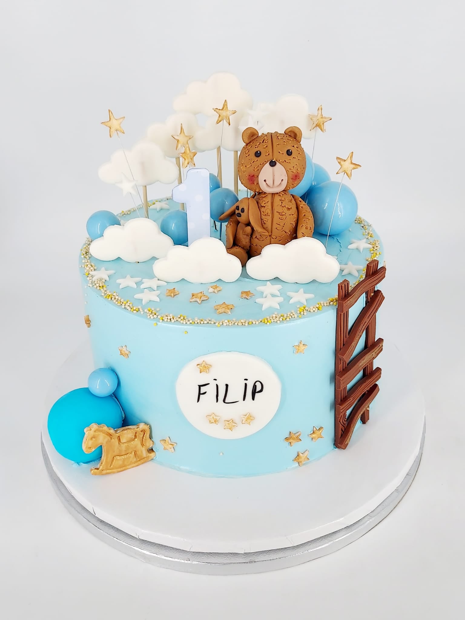 Tort pentru moț de băiat, de culoare albastră, personalizat cu numele Filip și decorat cu un ursuleț maro, cifra 1, norișori și steluțe aurii.
