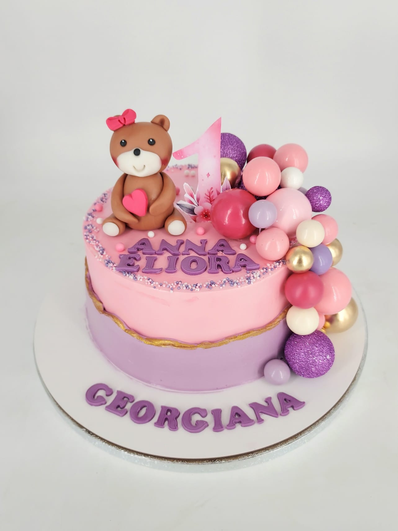 Tort pentru moț de fetiță, în culori roz și mov, personalizat cu numele Anna Eliora și decorat cu un ursuleț, cifra 1 și o cascadă de baloane colorate.