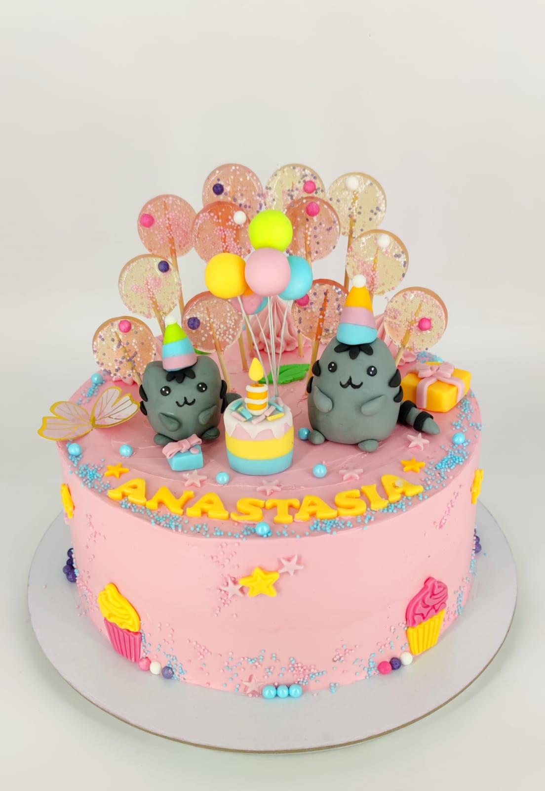 Tort de aniversare roz pentru copii, cu tematică Pusheen, decorat cu figurine pisici, acadele, baloane colorate și personalizat cu numele Anastasia.