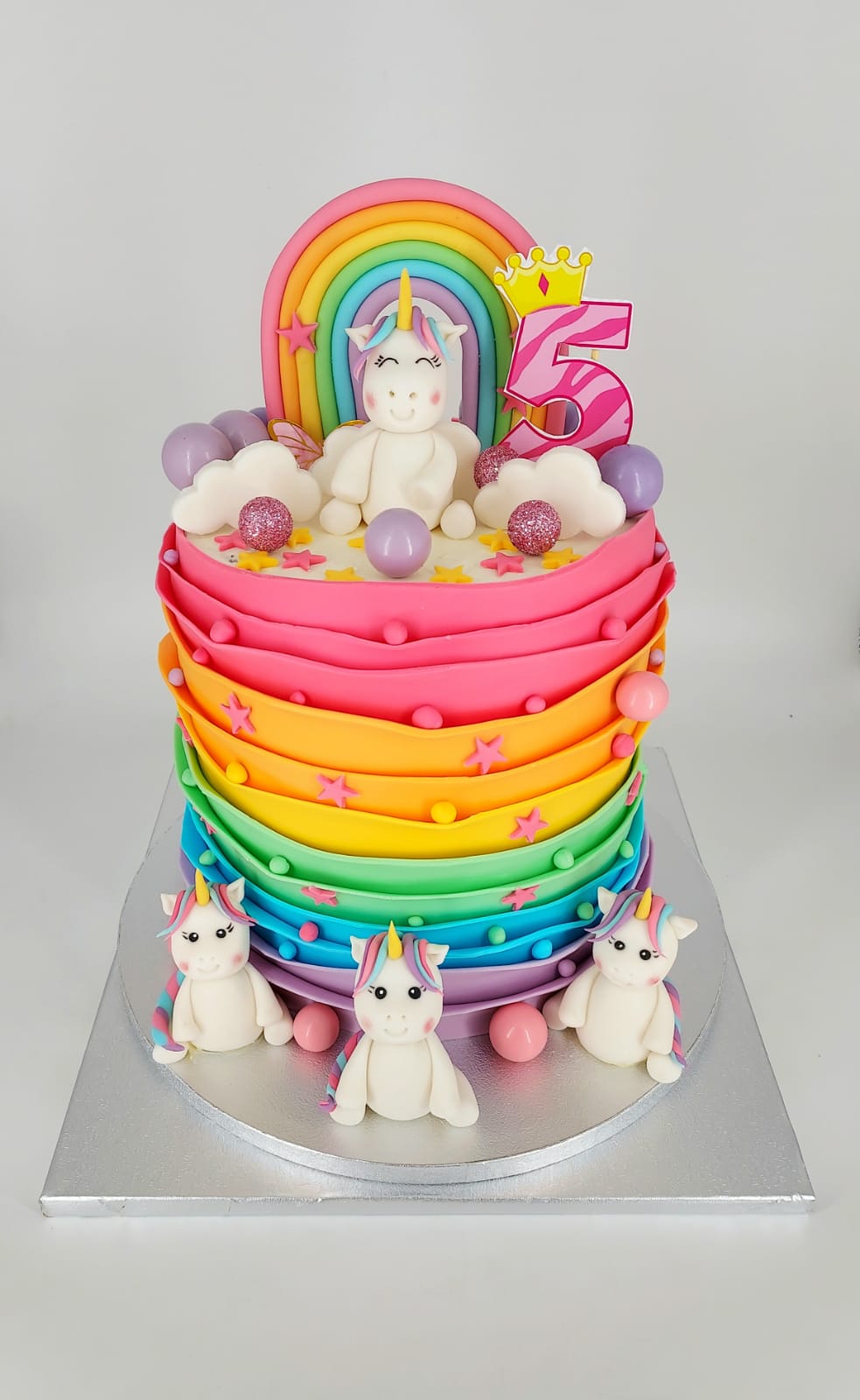 Tort aniversar înalt pentru fetițe, cu volane de cremă în culorile curcubeului, decorat cu un unicorn, cifra 5 și figurine din zahăr.