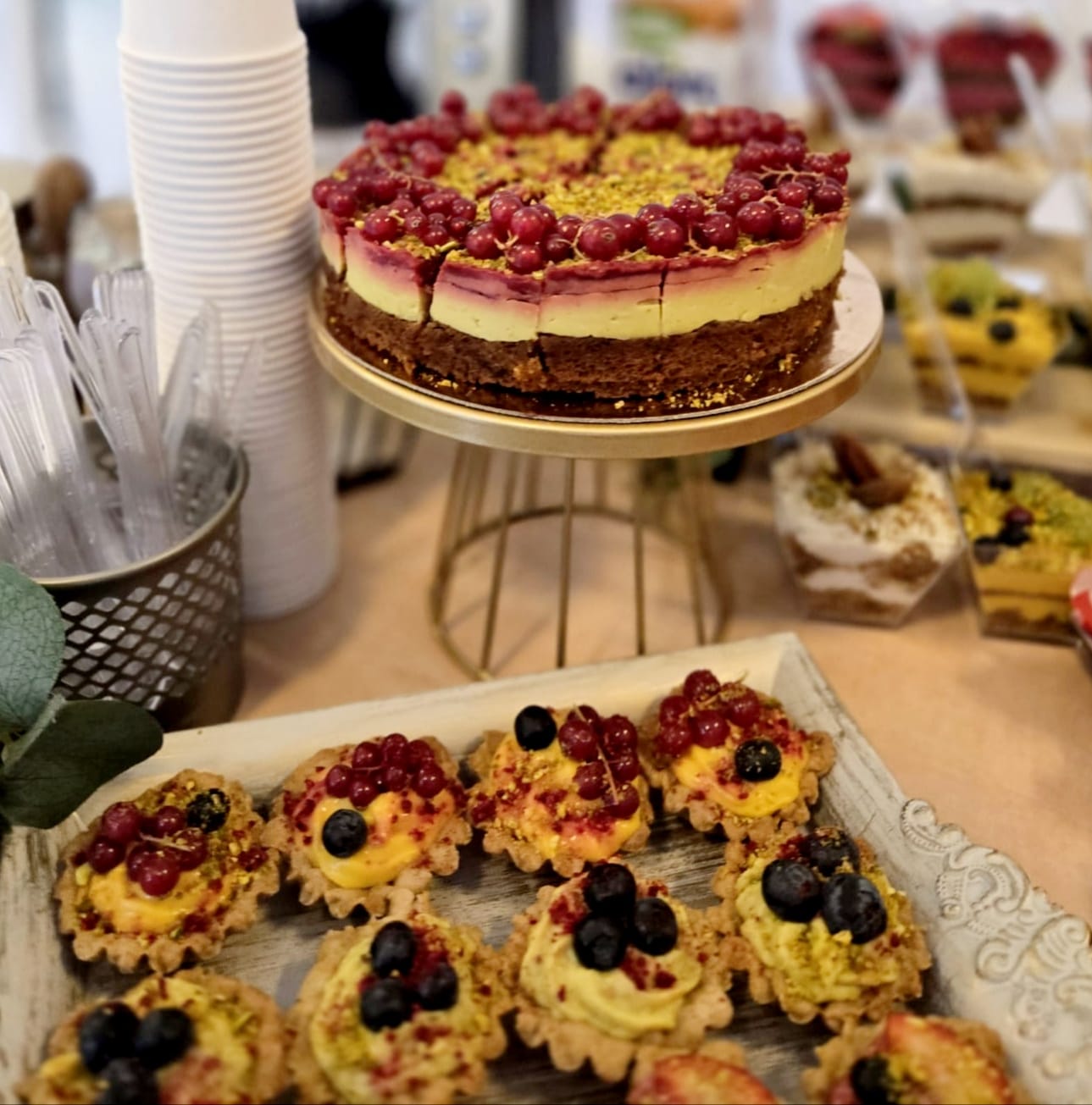 Tort vegan cu fistic și coacăze roșii pe un suport auriu, alături de mini tarte artizanale cu fructe, pregătite pentru un bufet de deserturi sănătoase la un eveniment.