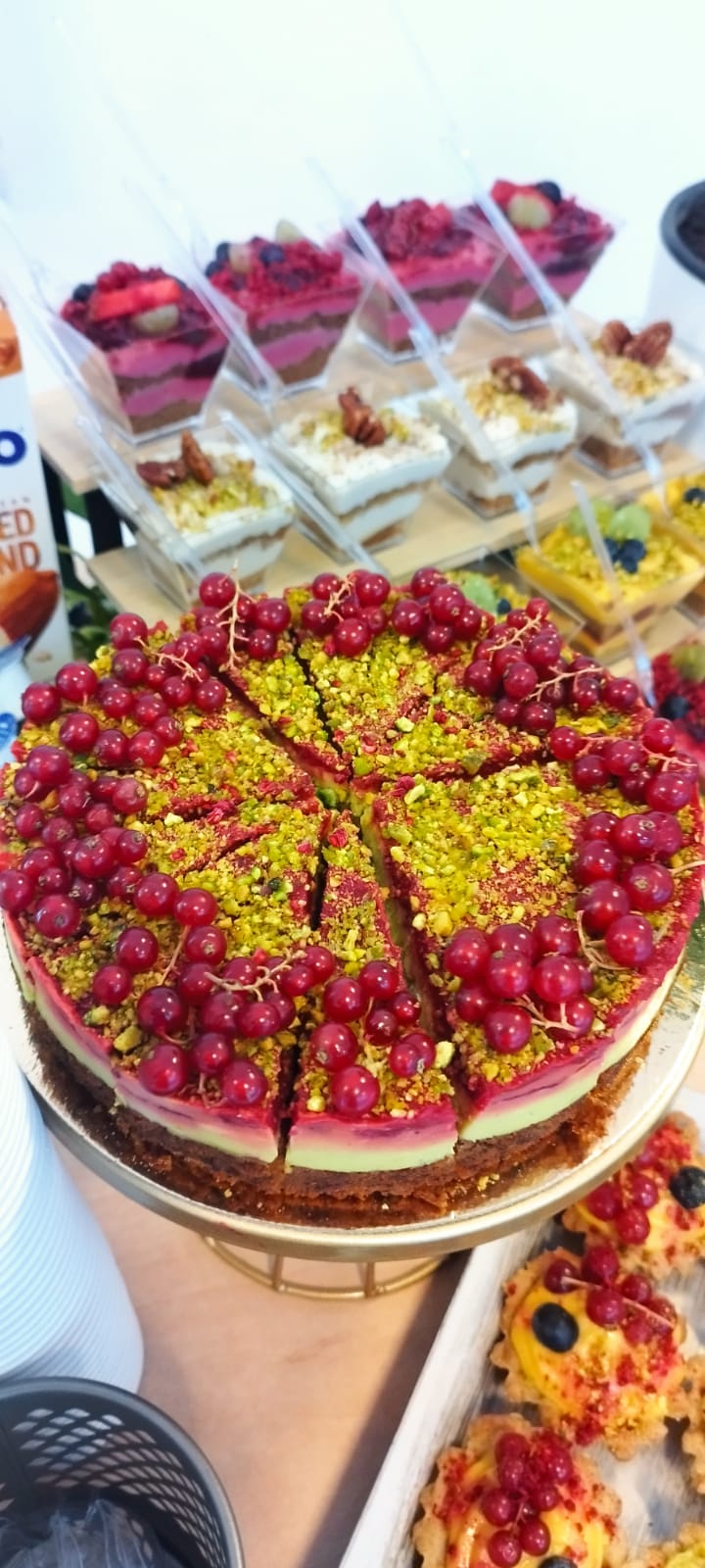 Vedere de sus a unui tort vegan artizanal decorat cu fistic și coacăze roșii, înconjurat de mini-deserturi asortate la pahar, într-un candy bar modern. Atelierul de limonada.