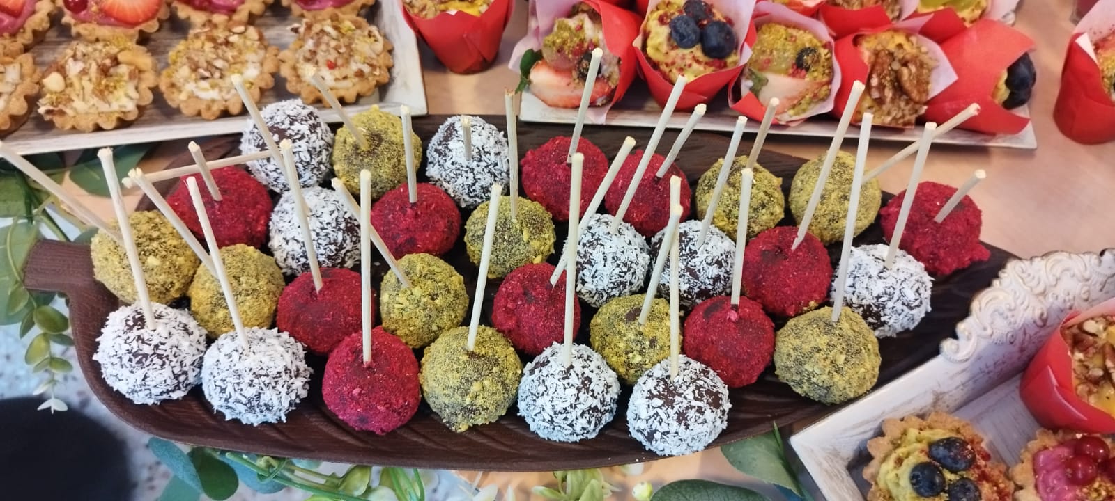Trufe raw-vegane pe băț (cake pops), acoperite cu fulgi de cocos, fistic și pudră de fructe roșii, așezate pe un platou în formă de frunză.